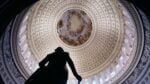 This image depicts inside the U.S. Capitol building. The ornate ceiling of the dome is shown, with a silhouette of a statue in the lower left portion of the image frame.