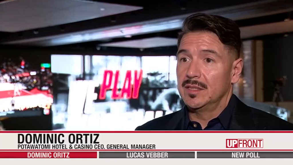 Photo of Potawatomi Casino & Hotel CEO Dominic Ortiz standing in front of a large LCD screen with the word "play" dispalyed on it.