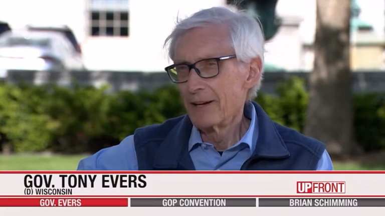This image shows Gov. Tony Evers sitting on a bench in front of the Wisconsin Capitol wearing a blue dress shirt and dark-gray fleece vest.