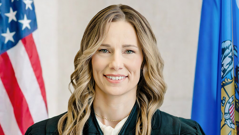 This image depicts Judge Lyndsey Brunette wearing judicial robes and flanked by the flags of the United States and the state of Wisconsin
