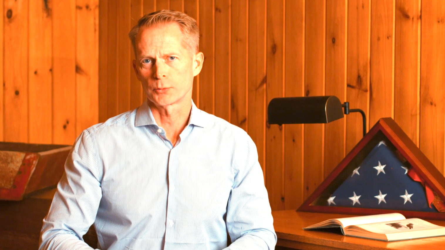 This image depicts GOP businessman and attorney Paul Wassgren seated in a room with walls of natural-finished vertical shiplap next to a table with an encased folded flag, an opened book and a black lamp upon it.